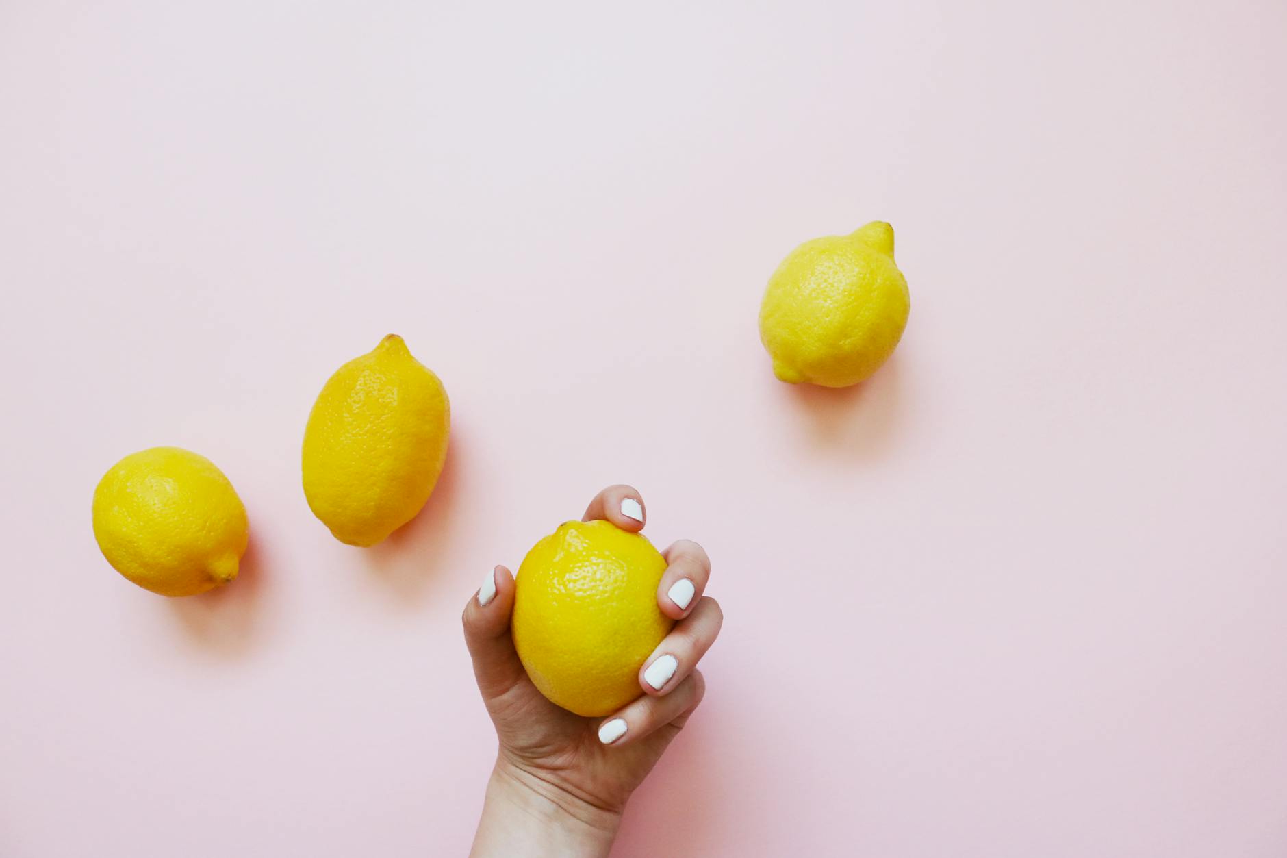 Hand holding a lemon on a soft pink background surrounded by additional lemons